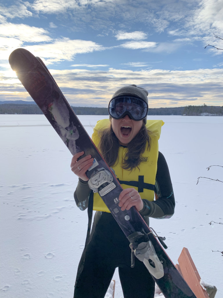 Photo of our very own store manager Julia wearing a wetsuit and life jacket in January standing on the dock with the frozen lake in the background. She's holding her snow skis, wearing her ski goggles. 